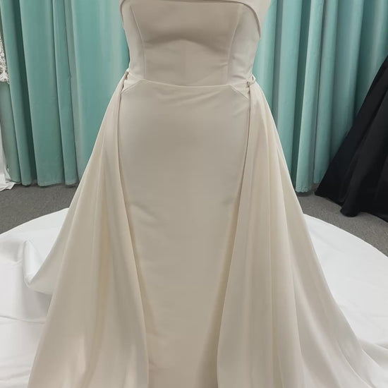 Oh Darling ivory strapless satin sheath wedding dress on mannequin with bandeau neckline, oversized statement bow at the waist, and voluminous overskirt.
