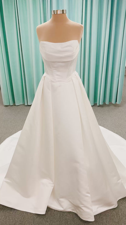 Stella York SY8084 ballgown wedding dress on mannequin with strapless ruched neckline, basque waist bodice, pleated satin skirt, and row of fabric-covered buttons