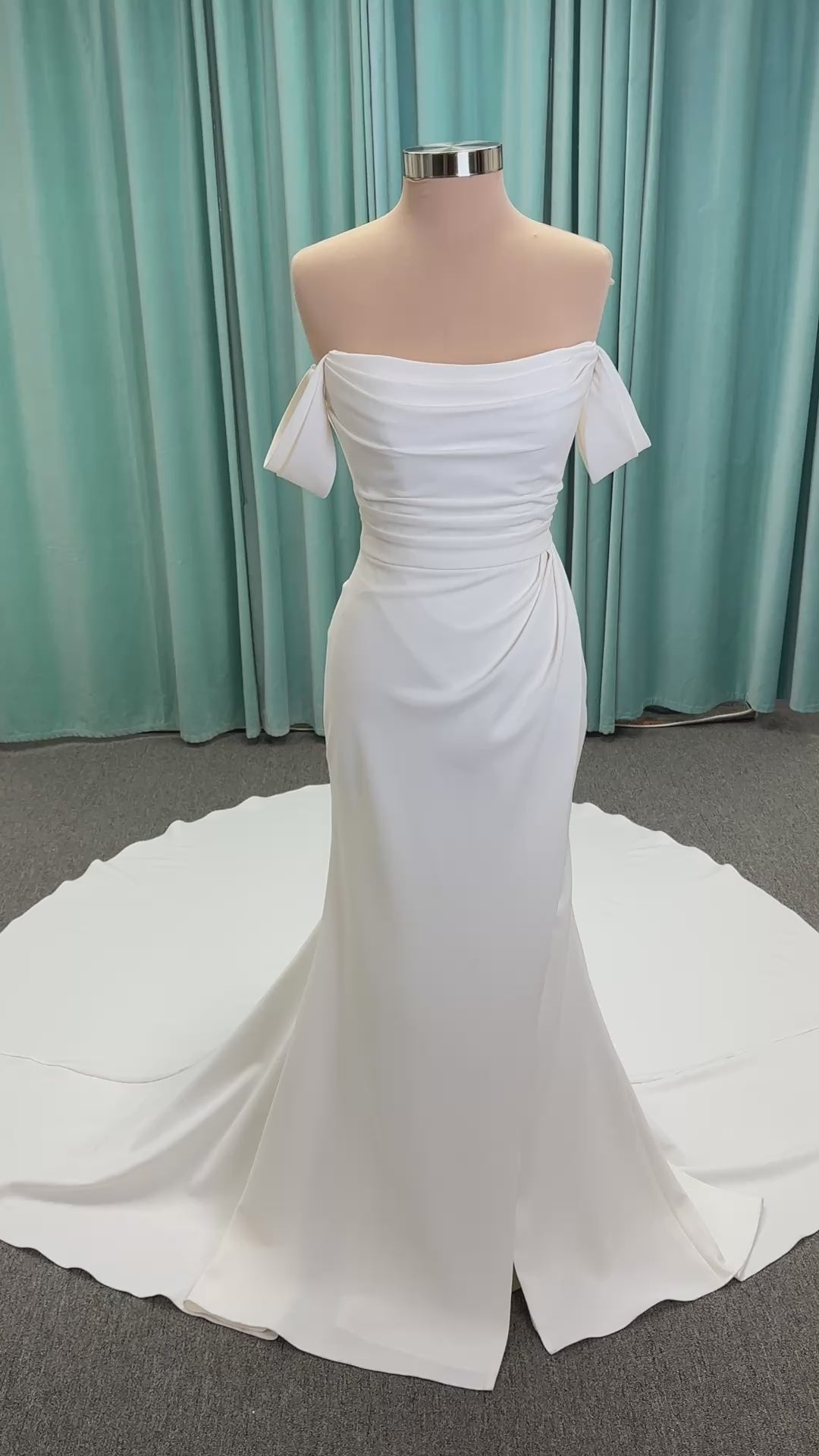 Tai ivory off-the-shoulder crepe sheath wedding dress on mannequin with draped bodice, front slit, and cathedral train.
